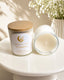 Candle labeled 'Sunshine' by 'Jou Collections' on a white surface with a vase of white flowers in the background.