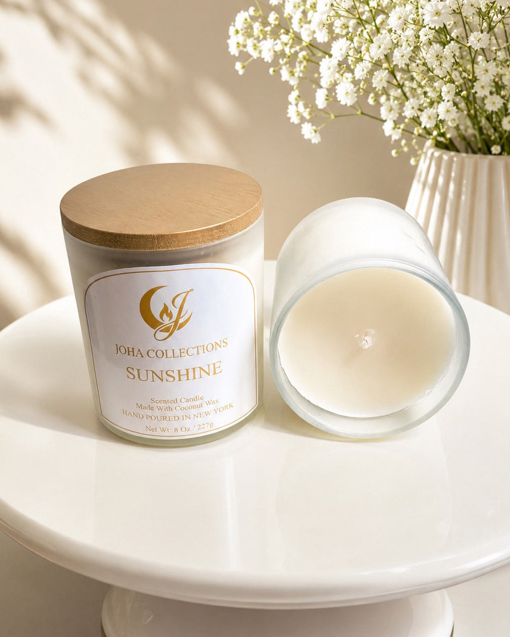 Candle labeled 'Sunshine' by 'Jou Collections' on a white surface with a vase of white flowers in the background. Main image