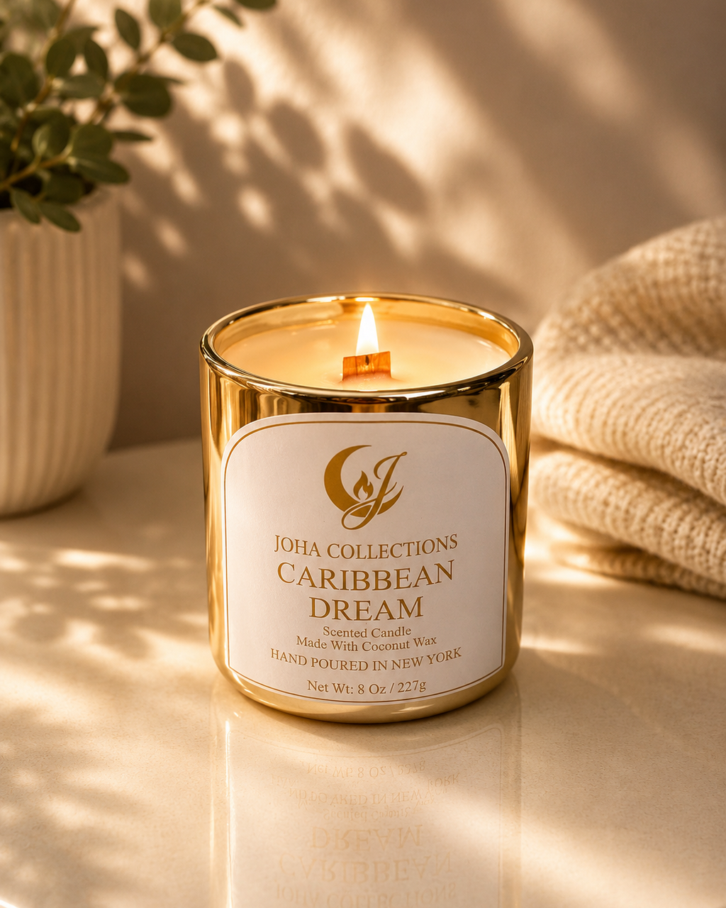 Candle with 'Joha Collections Caribbean Dream' label on a reflective surface with a plant and textured fabric in the background.