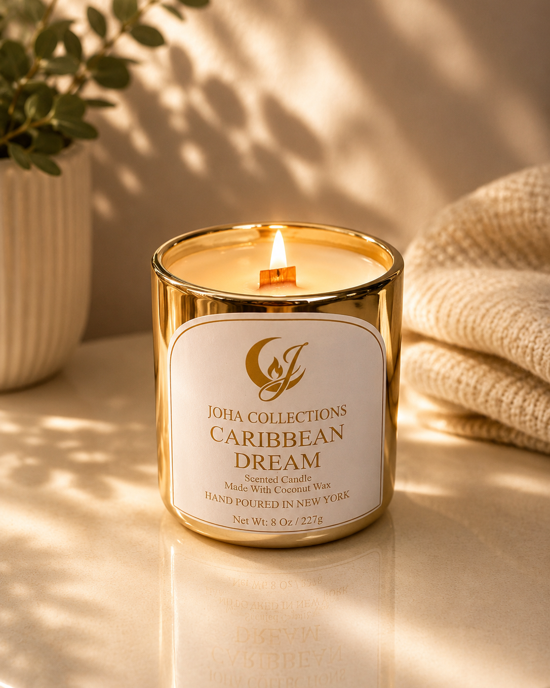 Candle with 'Joha Collections Caribbean Dream' label on a reflective surface with a plant and textured fabric in the background.