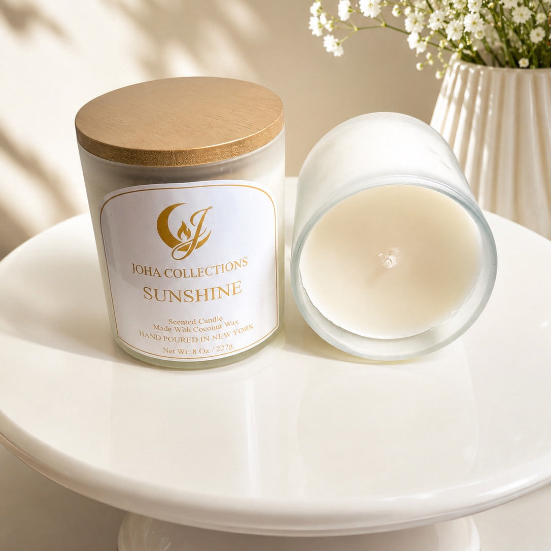 Candle labeled 'Sunshine' by 'Jou Collections' on a white surface with a vase of white flowers in the background.