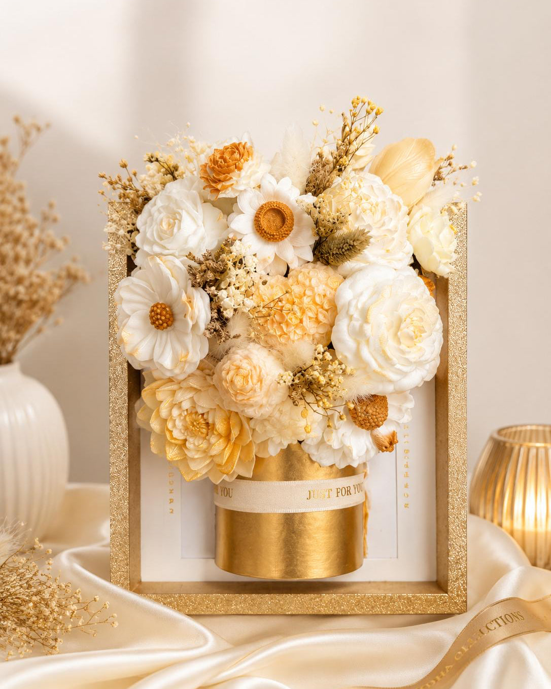 A decorative dried flower arrangement in a glittery gold box with a gold cylindrical base, styled on draped satin fabric with warm neutral props in the background. Main image